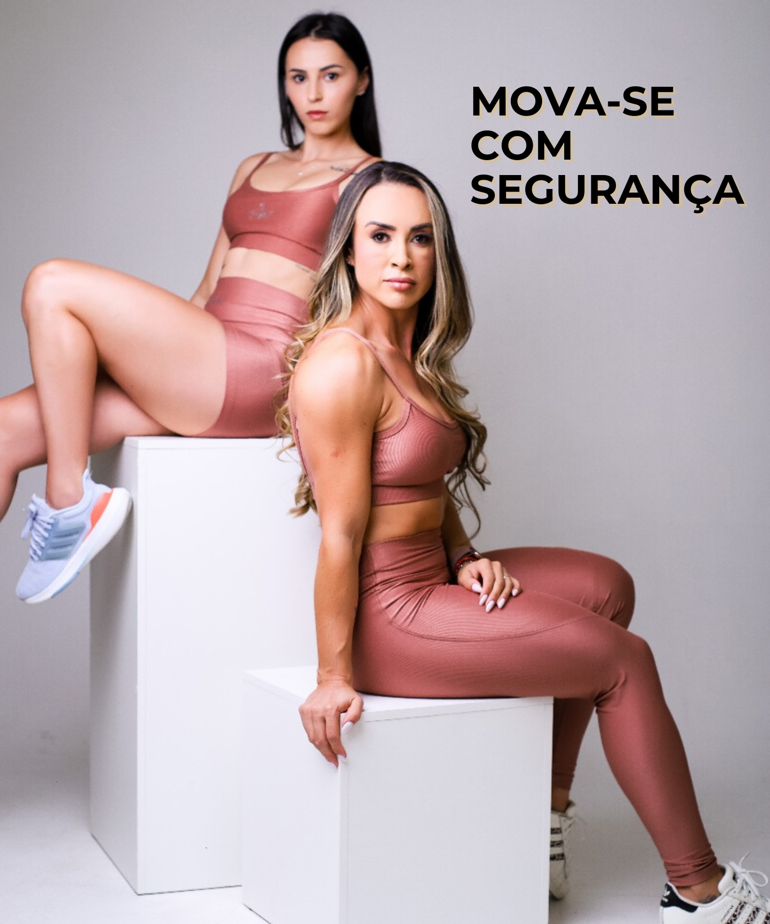 Duas mulheres em estúdio, usando conjuntos de moda fitness na cor terracota (top e leggings/shorts), em poses que transmitem confiança e conforto. Uma mulher loira olha diretamente para a câmera, enquanto a outra, de cabelos escuros, está ao fundo em pose relaxada. A imagem inclui o slogan 'MOVA-SE COM SEGURANÇA', destacando a qualidade e a segurança das roupas esportivas para um estilo de vida ativo e empoderado.
