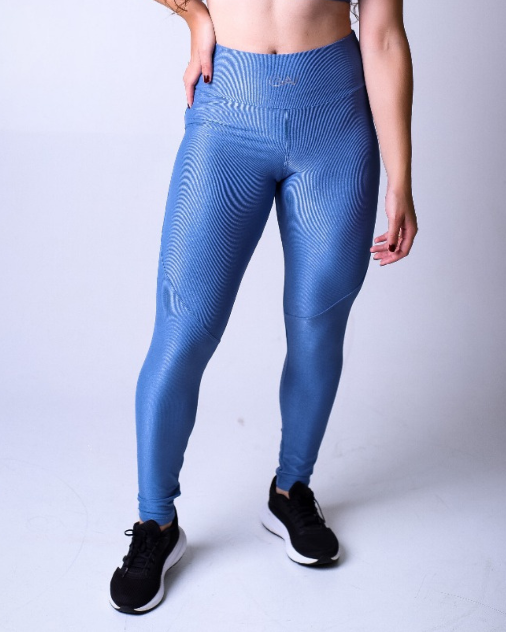 Mulher em pé, usando a mesma legging azul combinada com tênis preto. A iluminação destaca a textura do tecido e o cós alto com logo.
