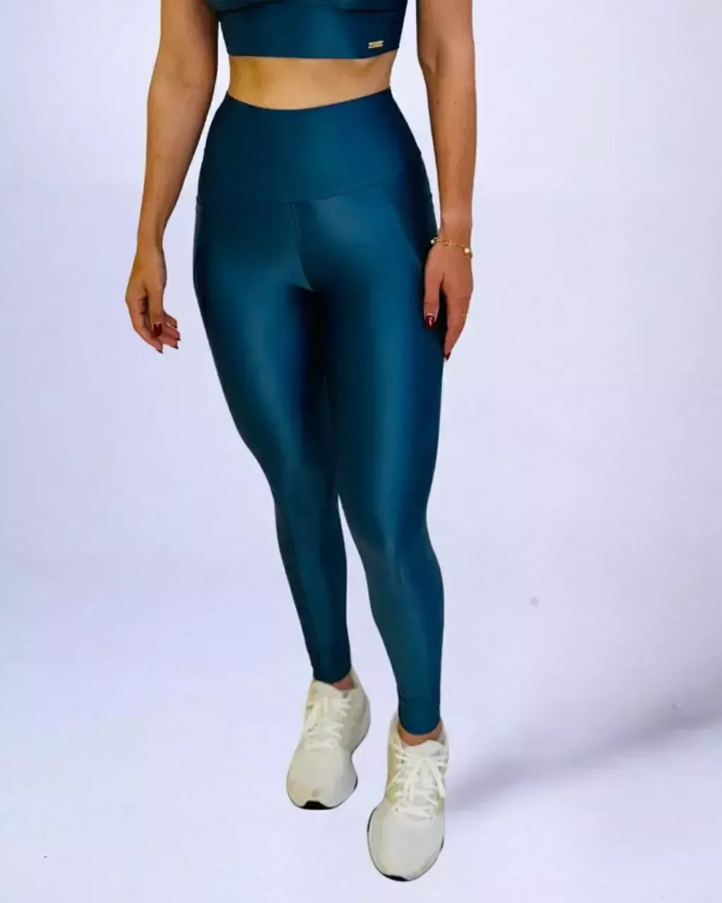 Mulher vestindo calça legging fitness de cintura alta azul-petróleo com brilho acetinado e tênis brancos. Um top esportivo da mesma cor é visível, completando o conjunto.