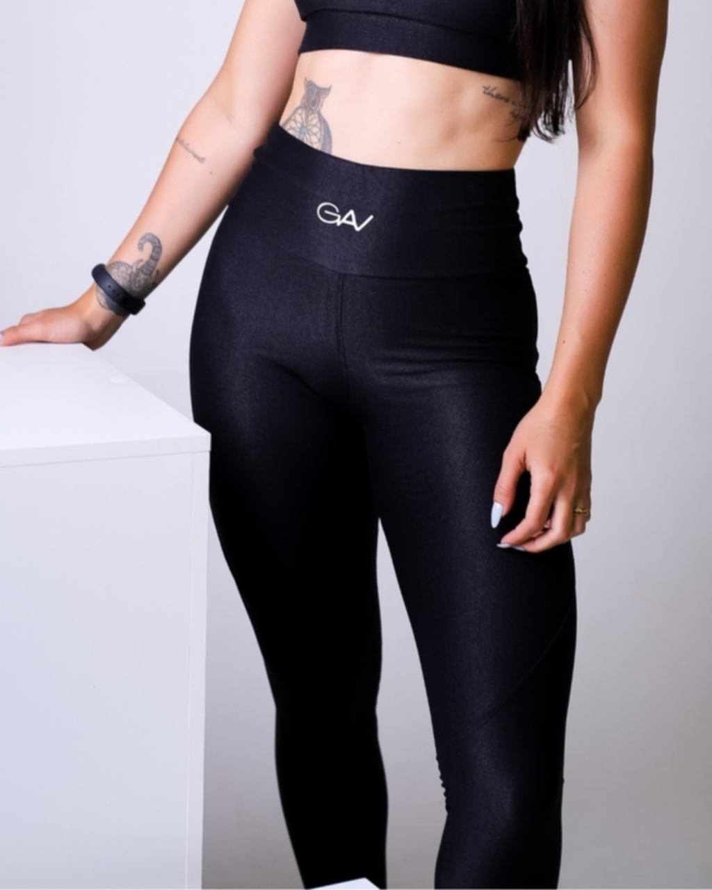 Mulher em pé, vestindo legging preta de cintura alta. O tecido tem leve brilho e o cós traz logo pequeno ao centro. O look é combinado com top preto.