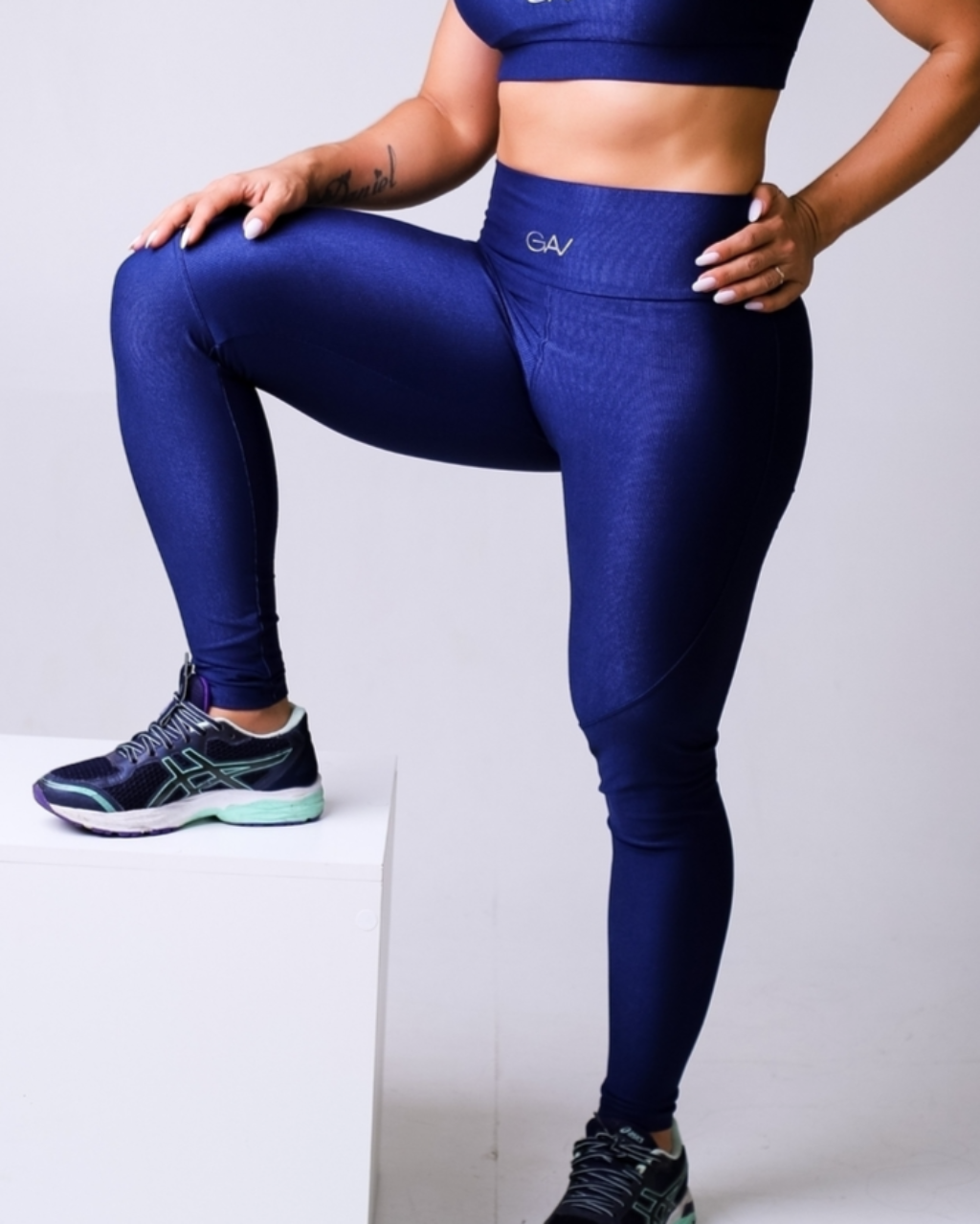 Mulher apoiando uma perna em um cubo branco, usando legging azul-marinho com tênis esportivo preto e detalhes verde-água. O foco está na modelagem firme e no cós alto.