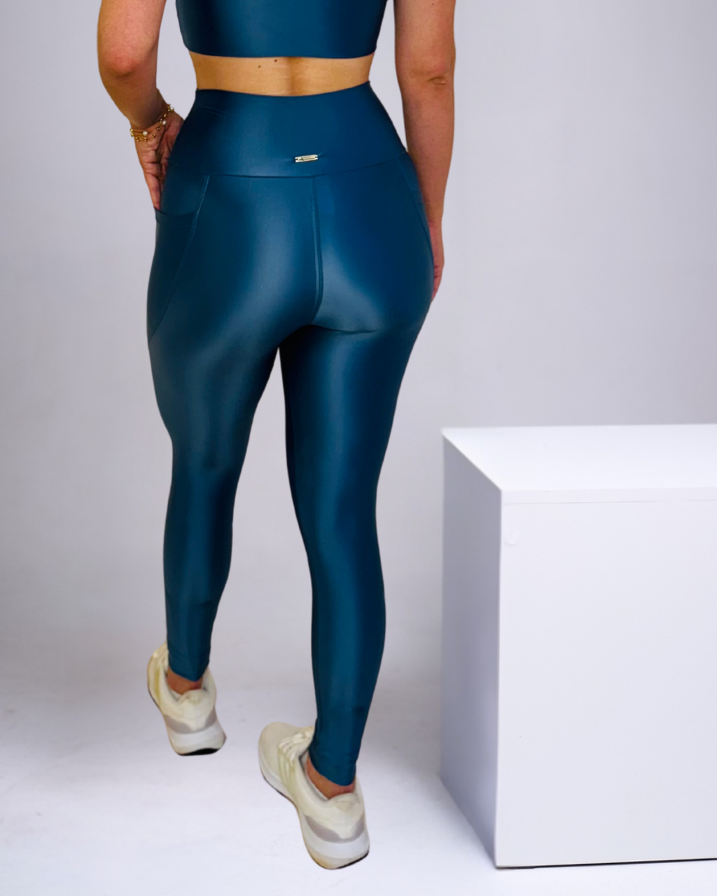 Vista traseira de uma pessoa usando um conjunto de academia completo na cor azul petróleo ou teal escuro. A calça legging é de cintura alta, com acabamento liso e levemente brilhante, e possui um pequeno bolso lateral visível na altura do quadril esquerdo. Um pequeno detalhe metálico (tag de marca) está visível na parte superior central do cós da legging. A pessoa também veste um top esportivo ou cropped da mesma cor, criando um visual coeso. Completa o look com tênis esportivos brancos com detalhes cinzas.