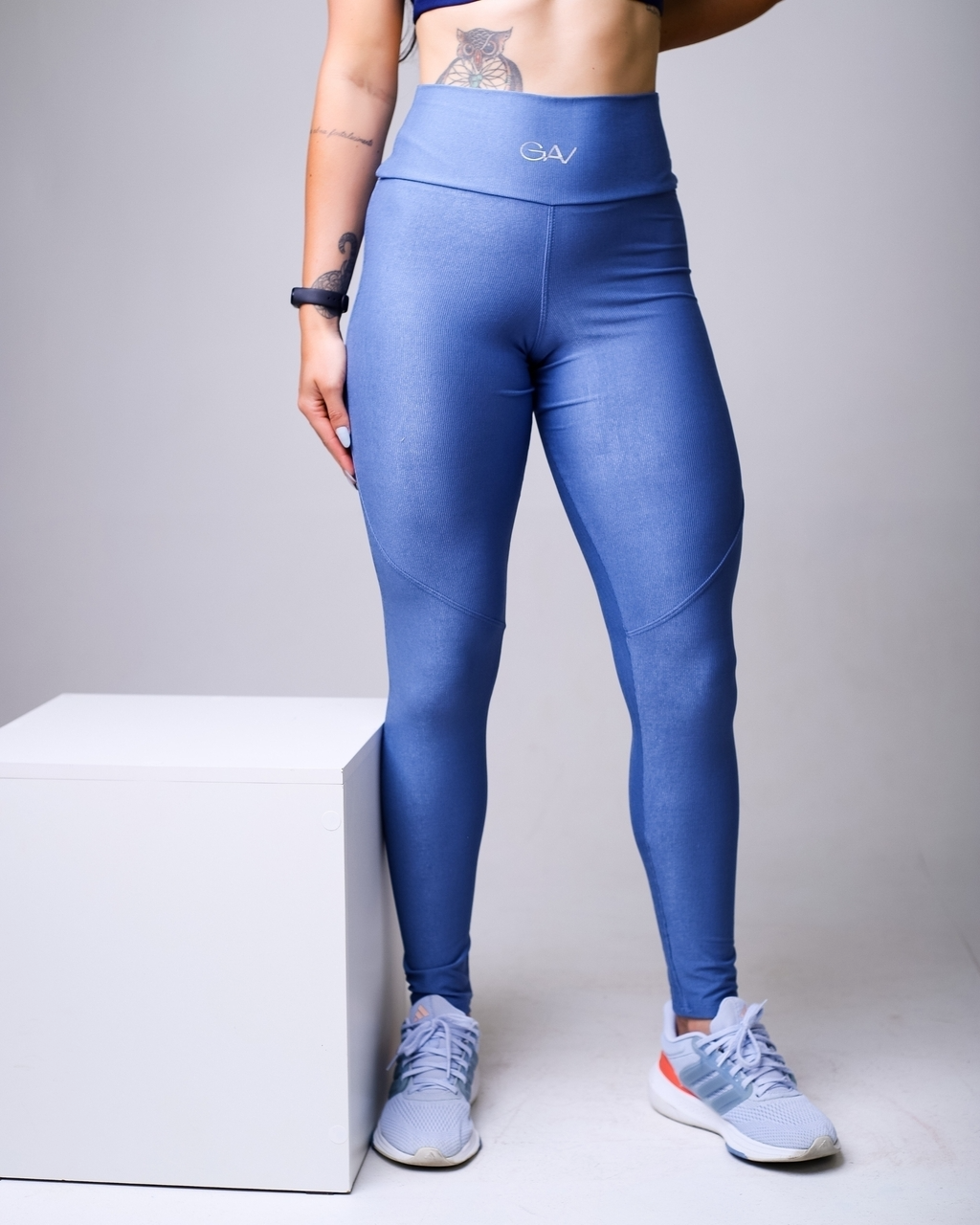 Mulher em pé, vestindo legging azul de cintura alta, combinada com tênis esportivo azul e branco. O foco está na frente da peça, mostrando o ajuste no corpo e o cós largo com logo discreto.